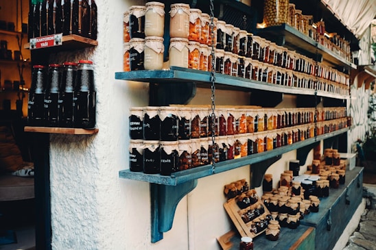 Shelves filled with a variety of jars containing preserves and sauces, labeled and neatly arranged. The jars are topped with brown paper and tied with string. There are different colors and types of preserves, creating a rustic and artisanal market display.