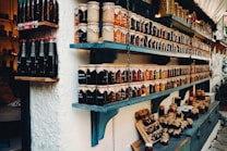 Shelves filled with a variety of jars containing preserves and sauces, labeled and neatly arranged. The jars are topped with brown paper and tied with string. There are different colors and types of preserves, creating a rustic and artisanal market display.