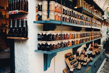 Shelves filled with a variety of jars containing preserves and sauces, labeled and neatly arranged. The jars are topped with brown paper and tied with string. There are different colors and types of preserves, creating a rustic and artisanal market display.