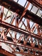 An intricate network of steel beams and girders forms a structural framework under a glass ceiling. The beams create intersecting patterns with a modern architectural design, allowing natural light to filter through. The surrounding environment includes reflections on the glass and shadows cast by the framework.