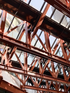 An intricate network of steel beams and girders forms a structural framework under a glass ceiling. The beams create intersecting patterns with a modern architectural design, allowing natural light to filter through. The surrounding environment includes reflections on the glass and shadows cast by the framework.
