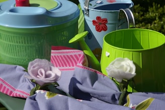 A collection of household items including a light blue plastic salad spinner, a green and light blue metal watering can with red floral designs, a green bucket, and an arrangement of pink and white felt flowers on a purple and pink striped cloth.