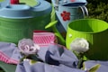 A collection of household items including a light blue plastic salad spinner, a green and light blue metal watering can with red floral designs, a green bucket, and an arrangement of pink and white felt flowers on a purple and pink striped cloth.
