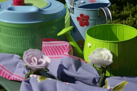 A collection of household items including a light blue plastic salad spinner, a green and light blue metal watering can with red floral designs, a green bucket, and an arrangement of pink and white felt flowers on a purple and pink striped cloth.