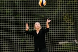 A person wearing black clothes is preparing to serve a volleyball. The background consists of a net with a blurred wooded area behind it.
