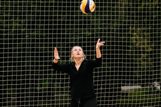 A person wearing black clothes is preparing to serve a volleyball. The background consists of a net with a blurred wooded area behind it.