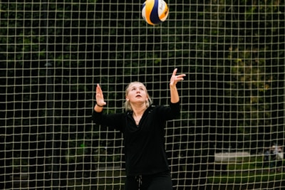 A person wearing black clothes is preparing to serve a volleyball. The background consists of a net with a blurred wooded area behind it.
