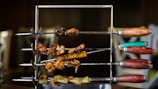 A set of skewers with various grilled meats arranged on a metal rack, featuring wooden handles of different colors such as red, green, and orange. The meats appear to be cooked, with crispy and charred bits visible.
