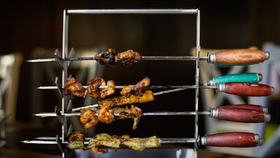 A set of skewers with various grilled meats arranged on a metal rack, featuring wooden handles of different colors such as red, green, and orange. The meats appear to be cooked, with crispy and charred bits visible.