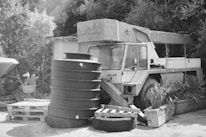 An old, dilapidated crane vehicle is situated in an outdoor area surrounded by foliage. It appears to be abandoned and is positioned next to a stack of large, cylindrical containers and wooden pallets. Various plants and bushes are growing nearby, adding to the rustic setting.