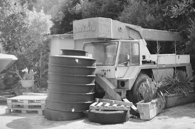 An old, dilapidated crane vehicle is situated in an outdoor area surrounded by foliage. It appears to be abandoned and is positioned next to a stack of large, cylindrical containers and wooden pallets. Various plants and bushes are growing nearby, adding to the rustic setting.