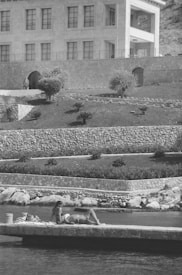 A woman in a bikini is sunbathing on a stone platform adjacent to the water. In the background, a large, stone building with multiple windows rises above a terraced landscape featuring neatly manicured shrubs and bushes. The scene is peaceful and serene, with a clear focus on relaxation and leisure.