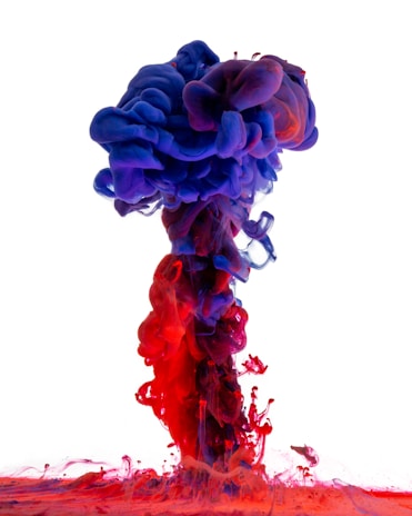 Vivid plumes of colorful ink swirl and mix in water, creating a striking gradient of reds and purples against a white background. The dense clouds of ink form a fluid, mushroom-like shape, showcasing the intricate interplay of light and color as they disperse into one another.