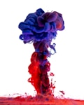 Vivid plumes of colorful ink swirl and mix in water, creating a striking gradient of reds and purples against a white background. The dense clouds of ink form a fluid, mushroom-like shape, showcasing the intricate interplay of light and color as they disperse into one another.