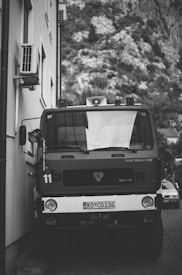 A vintage fire truck is parked closely alongside a building, with a mountainous landscape in the background. The vehicle has prominent headlights and a license plate on the front. Air conditioning units are installed on the wall of the adjacent building.