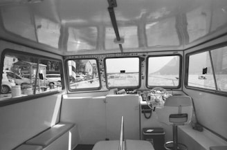 The interior of a boat's cabin with a steering wheel and control panel, surrounded by windows offering views of the surrounding waterfront, structures, and parked vehicles. The cabin has seating and a minimalistic setup.