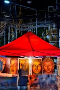 A vibrant street market at night features a red canopy under which several paintings are displayed. The paintings mainly depict various serene Buddha faces in warm tones. Electrical wires crisscross above, and the glow, likely from artificial lighting, bathes the scene, creating an intriguing interplay of light and shadow.