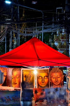 A vibrant street market at night features a red canopy under which several paintings are displayed. The paintings mainly depict various serene Buddha faces in warm tones. Electrical wires crisscross above, and the glow, likely from artificial lighting, bathes the scene, creating an intriguing interplay of light and shadow.