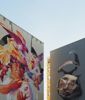 Two large murals adorn the sides of urban buildings. The left mural features a vibrant depiction of a person surrounded by colorful birds and whimsical elements like flowers and feathers. The right mural presents a more subdued, surreal image of a large face breaking through the surface, creating a three-dimensional effect.
