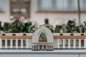 A scale model of a building with an arched central entrance and decorative windows. The structure features architectural details such as a brown roof and white walls. In the background, out-of-focus greenery suggests a natural surrounding.