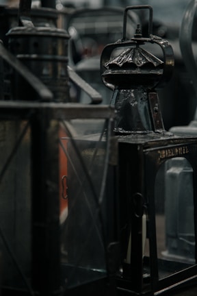 A collection of vintage lanterns with metallic and glass surfaces displayed closely together. The lighting casts a dim, muted ambiance, highlighting their aged texture with a focus on their intricate designs.