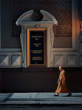 A person dressed in a saffron robe is walking along a dimly lit urban street, passing by a large ornate wall with a framed sign displaying the names Thomas Kail, Lin-Manuel Miranda, and Anthony Veneziale in gold lettering.