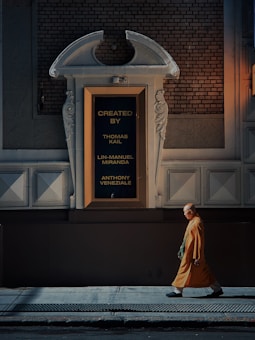 A person dressed in a saffron robe is walking along a dimly lit urban street, passing by a large ornate wall with a framed sign displaying the names Thomas Kail, Lin-Manuel Miranda, and Anthony Veneziale in gold lettering.