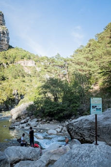 A peaceful riverside scene with three people situated near the water surrounded by large rock formations and lush greenery. One individual is shirtless and sitting on a rock, enjoying the serene environment. A posted sign indicates the presence of hiking or safety information. The backdrop of the scene includes rocky cliffs and a sky with few clouds, suggesting a natural and outdoor setting.