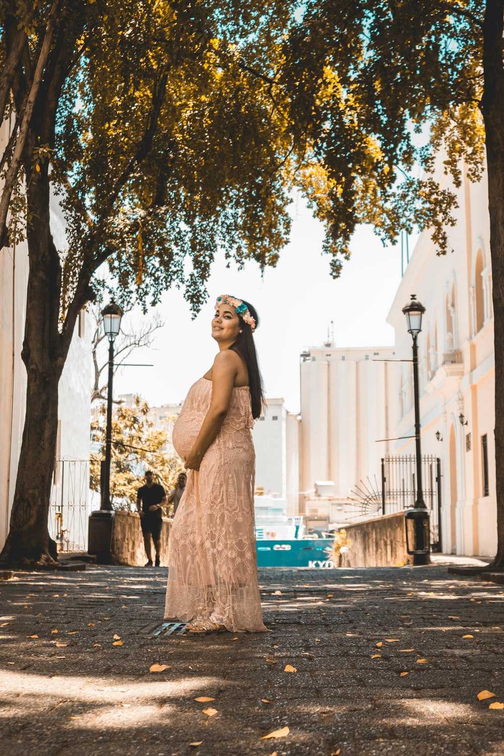 smiling pregnant woman wearing beige tube dress touching her tummy while standing on concrete pathway
