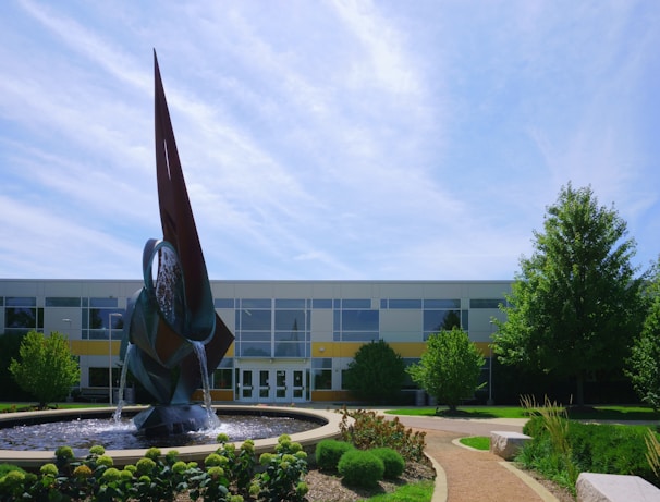 A modern building with large windows is set behind a prominent abstract metal sculpture that also functions as a fountain. Water cascades from the sculpture into a circular pool surrounded by neatly trimmed greenery. The scene is framed by a clear blue sky and various trees and shrubs, contributing to a serene outdoor setting.