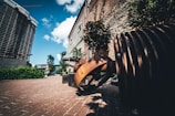 A piece of industrial art featuring large rusty metal gears on a paved pathway, surrounded by brick buildings and greenery. The sky is bright blue with a few scattered clouds, complementing the urban setting.