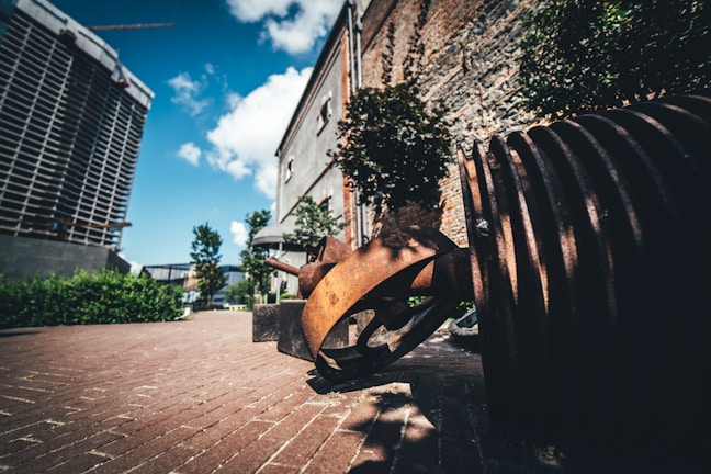 A piece of industrial art featuring large rusty metal gears on a paved pathway, surrounded by brick buildings and greenery. The sky is bright blue with a few scattered clouds, complementing the urban setting.