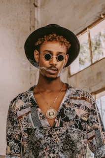A person wearing a patterned shirt and round sunglasses, accessorized with a large, ornate pendant necklace and a wide-brimmed hat. The setting appears to be an industrial or rustic space with large windows.