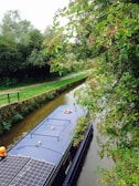 A narrowboat is traveling along a calm canal, bordered by lush green trees and vegetation. Solar panels are visible on the roof of the boat, along with a lifebuoy. Overhanging branches with berries create a peaceful and natural setting.