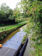 A narrowboat is traveling along a calm canal, bordered by lush green trees and vegetation. Solar panels are visible on the roof of the boat, along with a lifebuoy. Overhanging branches with berries create a peaceful and natural setting.