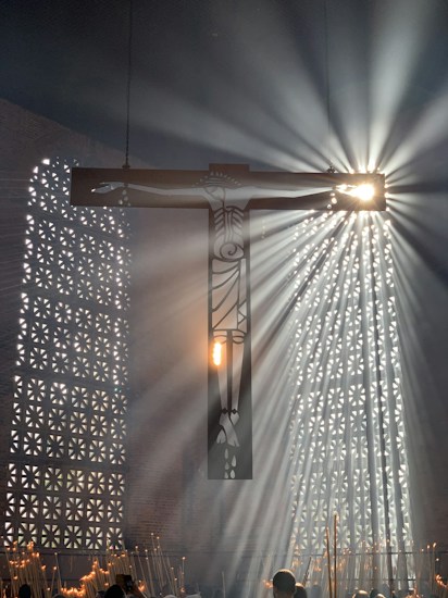 A large cross is suspended in a dimly lit interior space, with radiant beams of light streaming through a perforated wall. The light creates a dramatic effect as it shines around the silhouette of the cross, casting intricate shadows and illuminating the space. Several people are gathered below, holding candles that add to the ambient glow.