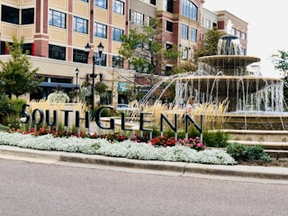 A large fountain is surrounded by colorful flowers and grasses with a modern multi-story building in the background. The area appears to be urban and well-maintained. There is a sign reading 'SOUTHGLENN' amidst the greenery.