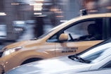 A yellow taxi is in motion on a city street, captured in a side profile with a driver visible inside. The background appears blurred, indicating movement and speed. The taxi has a sign on its side indicating independent ownership.