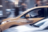 A yellow taxi is in motion on a city street, captured in a side profile with a driver visible inside. The background appears blurred, indicating movement and speed. The taxi has a sign on its side indicating independent ownership.