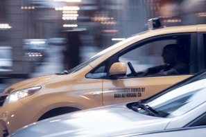 A yellow taxi is in motion on a city street, captured in a side profile with a driver visible inside. The background appears blurred, indicating movement and speed. The taxi has a sign on its side indicating independent ownership.