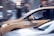 A yellow taxi is in motion on a city street, captured in a side profile with a driver visible inside. The background appears blurred, indicating movement and speed. The taxi has a sign on its side indicating independent ownership.
