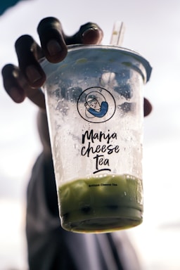 A hand holds a clear plastic cup with condensation on the outside. The cup is branded with 'Maria Cheese Tea' and contains a green beverage. The background is blurred, emphasizing the focus on the drink.