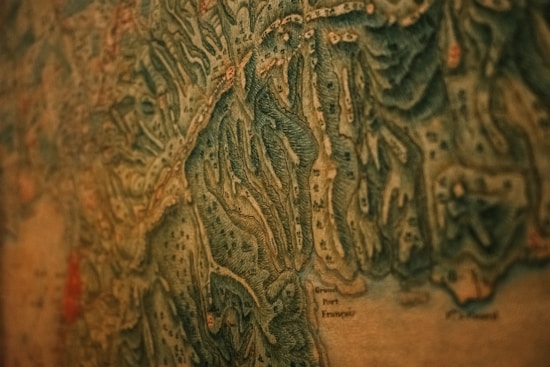 A close-up view of an old, detailed map with intricate topographical features. The map has various shades of green and brown, indicating different elevations and terrain types. Small markings and labels are scattered throughout, suggesting towns or landmarks.