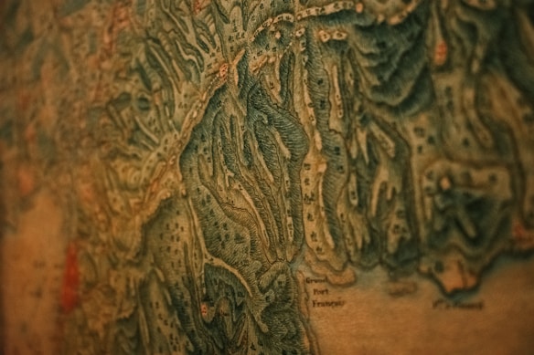 A close-up view of an old, detailed map with intricate topographical features. The map has various shades of green and brown, indicating different elevations and terrain types. Small markings and labels are scattered throughout, suggesting towns or landmarks.