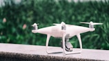 A white quadcopter drone is placed on a marble surface with a blurred background of green foliage. It features four propellers and a camera mounted on the underside.