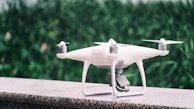 A white quadcopter drone is placed on a marble surface with a blurred background of green foliage. It features four propellers and a camera mounted on the underside.