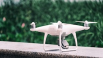 A white quadcopter drone is placed on a marble surface with a blurred background of green foliage. It features four propellers and a camera mounted on the underside.