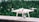 A white quadcopter drone is placed on a marble surface with a blurred background of green foliage. It features four propellers and a camera mounted on the underside.