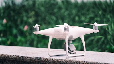 A white quadcopter drone is placed on a marble surface with a blurred background of green foliage. It features four propellers and a camera mounted on the underside.