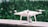 A white quadcopter drone is placed on a marble surface with a blurred background of green foliage. It features four propellers and a camera mounted on the underside.